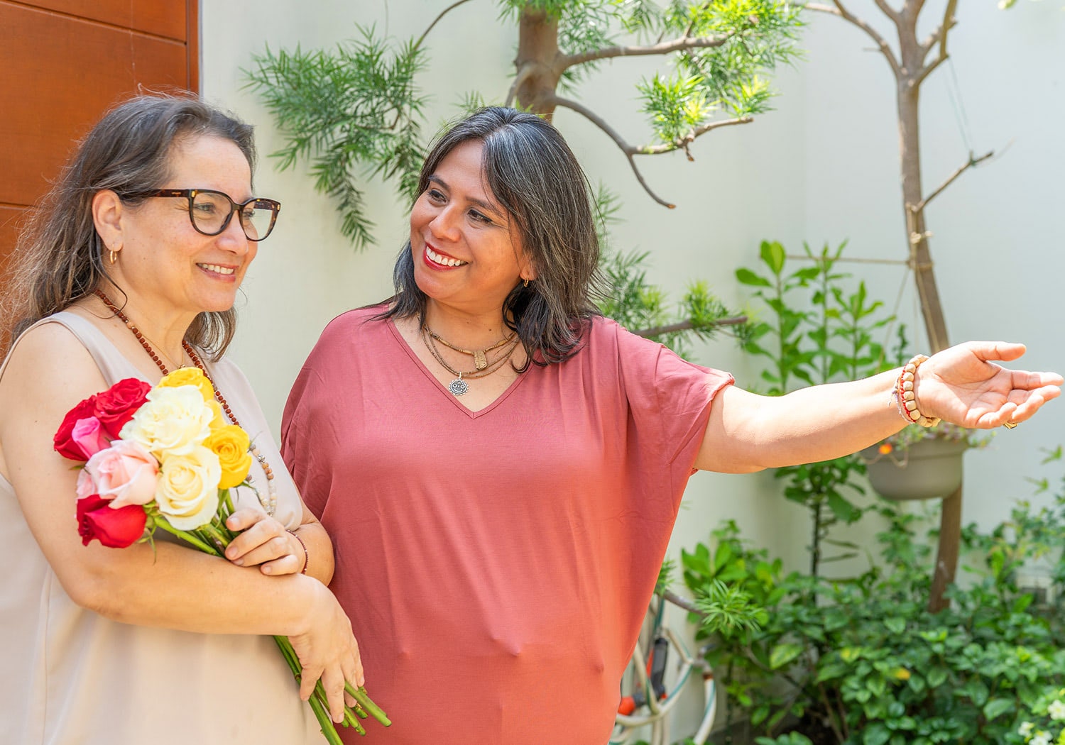 Two sisters smiling in a garden, one holding a colorful bouquet of roses.